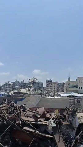 The video shows a wide shot of a city skyline under a clear blue sky with some white clouds. The foreground is filled with a pile of rubble and debris, including twisted metal and what appears to be broken building materials. Behind the debris, the middle ground of the image reveals a heavily damaged urban landscape. Several buildings are in various states of destruction, with collapsed roofs, broken windows, and exposed structural elements. One building in particular, on the left side of the frame, appears to be a multi-story concrete structure that has partially collapsed and is severely damaged, with some floors still standing but in a precarious state. The overall scene suggests a recent destructive event, such as an explosion or conflict, that has devastated a part of the city. No people are visible in the frame. The camera appears to be static or moving very slowly.