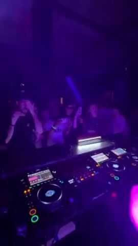 A DJ works at an electronic music console, lit by purple lights, in front of a crowd of dancing people. The DJ console is black, with four circular, metallic controls, four touchscreen displays, and an array of buttons, switches, and sliders. The DJ wears a neon yellow top. The crowd is standing in front of the DJ, with their arms raised. Some people are holding up their phones, presumably to record the performance. The environment appears to be a nightclub, with a dark interior and exposed beams on the ceiling. The mood is energetic, and the music is loud.