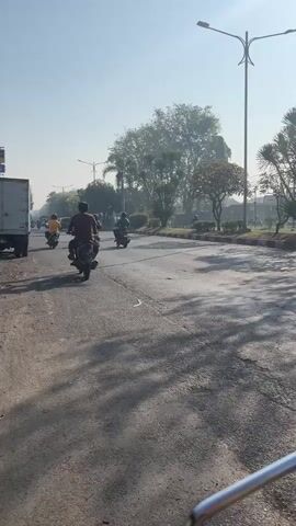 The video captures a street scene with several vehicles, including motorcycles and a white van. The weather appears clear and sunny, with shadows cast on the asphalt road. In the foreground, the handlebars of a motorcycle are visible, suggesting the footage is from the perspective of a rider. Several individuals on motorcycles are seen moving along the road. The environment includes trees and streetlights, indicating an urban or suburban setting. The overall atmosphere is that of daily traffic.