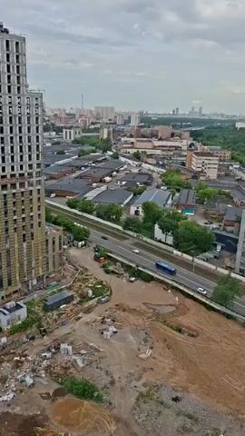 The video shows an aerial view of a construction site and surrounding industrial area. In the foreground, a tall, unfinished building is under construction, with scaffolding and building materials visible. The ground around the building is disturbed earth and debris. A road runs through the middle of the scene, with several vehicles, including trucks and a bus, traveling on it. Beyond the road, there are many low-rise industrial buildings with dark roofs and some green trees. In the distance, a cityscape with taller buildings is visible under a cloudy sky.