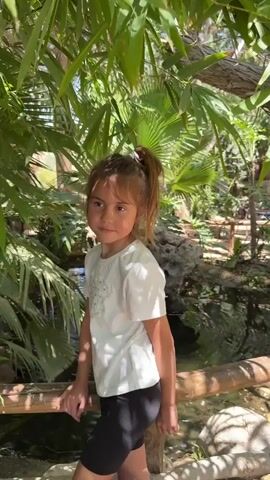 A young girl with brown hair in a ponytail, wearing a white t-shirt and black shorts, stands by a wooden railing in an outdoor setting. She is looking off to the side with a neutral expression. Behind her is a body of water, possibly a stream or pond, with a small waterfall cascading over rocks. Lush green foliage, including palm leaves, surrounds the area, suggesting a tropical or garden environment. The lighting indicates it is daytime, with sunlight filtering through the leaves.