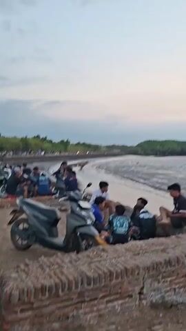 The video opens on a group of young males gathered near a body of water, possibly a mangrove forest, during what appears to be twilight. They are seated on a low brick wall, some engaged in conversation, others looking out towards the water. Several motorcycles are parked nearby, indicating a mode of transportation. The ground is a mix of dirt and gravel, with the brick wall forming a border. As the video progresses, the camera pans, revealing more people and motorcycles in the background, suggesting a social gathering or a popular spot. The water's edge is visible, with some muddy areas and ripples on the surface. The sky is overcast with soft hues of pink and grey, contributing to the tranquil atmosphere. The camera continues to pan and zoom out, showing a wider view of the coastline, the mangrove trees, and a broader expanse of people and vehicles. The overall scene depicts a casual evening gathering of people by the water, with motorcycles being a prominent feature.