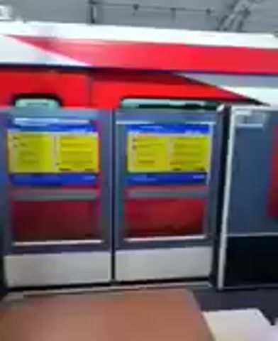 The video shows a view of a train station platform. In the foreground, a brown counter is visible. Behind the counter, there are two ticket machines with yellow screens displaying information. To the right of the ticket machines, a person wearing a white top and pink pants is standing, interacting with one of the ticket machines. Further down the platform, more people are visible, some standing and some walking. A train is docked at the platform, with its doors open. The station appears to be covered by a roof. Above the platform, a red and white sign is mounted on the ceiling.