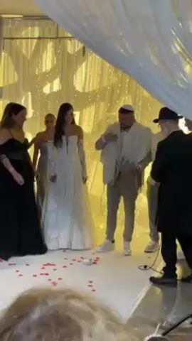 A medium shot of a groom at a Jewish wedding, stomping on a glass, breaking it. The groom wears a grey suit, white sneakers, a white knit kippah on his head, and a white shawl with tassels. A bride stands to his left, wearing a long, light dress with matching sleeves. Several other people stand on either side of them, a woman in a black dress, a young woman in a silver dress, and two young men in dress shirts and pants. A man in a suit stands to the far right, holding a paper. They stand on a platform covered in white fabric and scattered with red rose petals. Above them, white fabric is arranged in a canopy. Behind them, a yellow light installation is displayed. At the start of the shot, the groom is getting situated for the glass breaking. A woman to the groom’s left helps the groom get ready to break the glass. After breaking it, the people around the groom cheer, clap their hands and raise their hands.