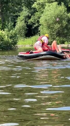Two people are on an inflatable raft in a body of water. One person is kneeling and appears to be assisting the other person, who is lying down. Both individuals are wearing life vests. The raft is black and red. The water is calm with ripples. The background consists of green trees and foliage along the bank of the water. The sky is visible and appears clear.