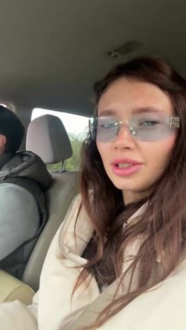 A young woman wearing pale-blue sunglasses and a light-beige jacket films a selfie video inside a car. She has long brown hair and is mouthing the words to the song playing on the car radio while making hand gestures. A man wearing glasses and a grey vest is in the passenger seat. He makes hand gestures and moves to the music as well, smiling. The vehicle is in motion.