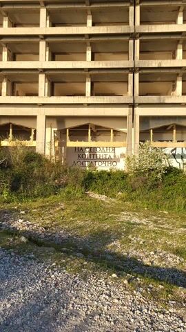 The video shows a close-up view of a large, unfinished concrete building, possibly an abandoned hotel or commercial structure. The building has multiple rectangular openings where windows would typically be, but the glass is missing or broken. The facade is weathered and stained. In the foreground, overgrown vegetation and a gravelly patch of ground cover the area in front of the building.

A sign is visible on one of the building's levels, painted in what appears to be Russian: "НА СЕГОДНЯ КОНТЕНТА ДОСТАТОЧНО" followed by the number 4308. The sign is integrated into the building's architecture, appearing as part of its design or graffiti. The sunlight casts shadows on the building and the vegetation, suggesting it is daytime.
