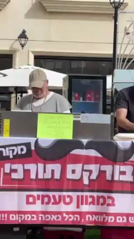The video shows two men standing behind a street vendor booth. The booth is advertising Turkish burekas, with a sign in Hebrew featuring a large mustache graphic. The older man on the left is wearing a tan baseball cap and a gray shirt, focused on his work. The younger man on the right, sporting a beard and a black t-shirt, is looking off-camera and appears to be interacting with someone not visible. A small refrigerator stocked with beverages is behind them. The scene is bright, suggesting daytime, and the setting appears to be an outdoor market or pedestrian zone with building facades in the background.
