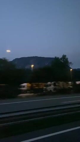 The video shows a view from a moving vehicle at dusk or night. A highway with lane markings is visible in the foreground. Beyond the highway, there are trees and a low wall or barrier. In the distance, a mountain range is silhouetted against a dark blue sky. Some lights, possibly streetlights or vehicle headlights, are visible among the trees and in the distance. There are also some stationary vehicles, possibly trucks or RVs, parked near the highway. The overall impression is of a twilight journey along a rural or semi-rural highway.