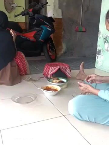 The video shows a group of people sitting on a tiled floor, eating a meal. There is a motorcycle parked in the background, as well as a broom leaning against a wall. Food is served in bowls and on plates. One person is wearing a blue hijab, and another is wearing a light blue dress. The overall scene appears to be informal and domestic.