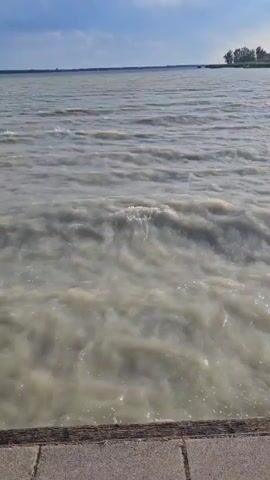 The video shows a body of water, likely a lake or river, with choppy, light-colored waves. The water appears somewhat murky. In the background, a distant shoreline is visible with trees and vegetation. The sky is partly cloudy with patches of blue. The foreground shows a textured surface, possibly a wooden or stone walkway, with a corner of a concrete structure visible on the right. No people or animals are visible. The overall impression is an outdoor scene with a focus on the movement of the water under a daytime sky.
