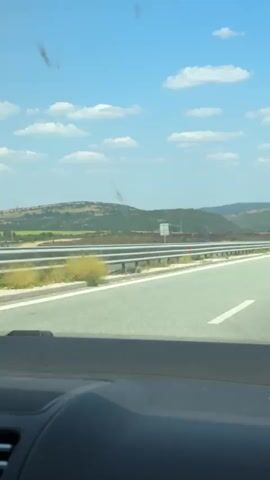 The video shows a view from inside a vehicle driving on a highway. The highway is paved and has white lane markings. A metal guardrail runs alongside the road. In the distance, there are rolling hills covered in green vegetation and some patches of darker, possibly burned areas. The sky is blue with scattered white clouds. A green bus is visible driving in the opposite direction on the highway. There are no people visible outside the vehicle.