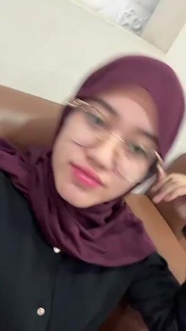 A close-up, selfie-style video shows a young woman indoors. She is wearing a dark maroon hijab and a black top. Her glasses have gold-colored frames, and she has bright pink lipstick on. She also wears a ring on her right hand.

Throughout the video, she is looking at the camera. She initially adjusts her hijab with her right hand. Around the 0:02 mark, she moves her head slightly to the side, looking directly at the camera, and then smiles, revealing braces on her teeth. The background is simple, with light-colored walls and a glimpse of a brown surface, possibly a chair.