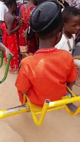 A young boy, dressed in a bright orange outfit and a black cap, sits on a yellow seesaw. He has a red beaded necklace around his neck. The seesaw is on a sandy surface, likely a playground.  Behind him, there is a group of people, some in colorful, traditional clothing. There is a faint musical sound in the background. The camera is handheld and stays mostly still, focusing on the boy. The scene appears to be taking place outdoors, possibly at a celebration or gathering.