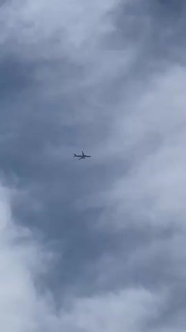 A silhouette of an airplane is visible in the sky, high above the clouds. The clouds are a mixture of dark and light gray, creating a dramatic backdrop for the flying object. The airplane appears to be moving across the frame from left to right. The video is shot from below, looking up at the sky. There is no audio.