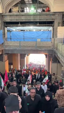 The video shows a crowd of people walking down a set of stairs and through an arched opening. Many of the people are wearing black clothing. Several individuals are holding flags, including flags with the colors red, white, and green, and some flags with Arabic text. The environment appears to be an indoor structure with concrete walls and a raised walkway or platform above. The lighting suggests it is daytime. The people are moving in a single direction, descending the stairs.