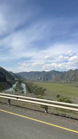 A video captures a scenic drive through a mountainous region with a river. The camera pans across a wide, green valley flanked by imposing mountains under a bright blue sky with scattered clouds. A calm, turquoise river winds through the valley. A road, with a white guardrail and a yellow line, runs along the edge of the frame, indicating the perspective of a moving vehicle. In the distance, a few people are visible standing near a parked black SUV with a roof carrier, suggesting a brief stop to admire the view. Further ahead, a few traffic signs are visible near the roadside, including a red and blue circular sign with a diagonal red bar and a triangular sign with a depiction of falling rocks, warning of potential hazards. The overall impression is one of natural beauty and a journey through a remote landscape.