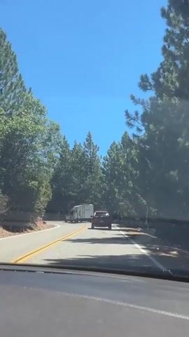 The video shows a view from inside a vehicle driving on a paved road. A red car is in front of the vehicle, also driving on the road. To the left of the red car and further up the road is a white trailer being towed by another vehicle. The road is surrounded by tall green pine trees and a clear blue sky. The road curves to the right.