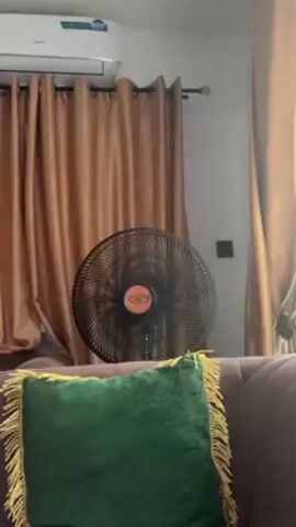 The video shows an indoor scene. In the foreground, there's a plush, emerald green square pillow with gold fringe resting on a dark, velvet-like sofa. Behind the pillow stands a black standing fan with an orange center, its blades spinning. In the background, beige curtains hang from a rod, slightly concealing the wall behind them. Above the curtains, an air conditioning unit is mounted on the wall. A small, dark square is also visible on the wall next to the curtains. The lighting is soft and diffused, suggesting daylight.