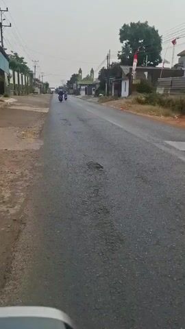 A low-angle, first-person perspective shot captures a view down a paved, two-lane road. The road is in poor condition, with multiple potholes visible. The sky is overcast. On either side of the road are small buildings, some with residential appearances and others possibly businesses. A green-domed building, possibly a mosque, is visible in the distance. A person on a motorcycle is traveling away from the camera in the left lane. Another person on a motorcycle is further ahead, also traveling away from the camera. The overall environment suggests a rural or suburban setting in a tropical climate, given the presence of lush trees and the style of the buildings. There are power lines overhead. A red and white Indonesian flag is attached to a pole on the right side of the road.