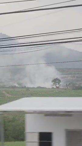 The video depicts a fire burning in a field. White smoke rises and merges with the overcast sky. The field is green, with some trees and bushes visible. In the background, a low mountain range is partially obscured by the haze. Utility lines are visible in the foreground, partially obscuring the view. The camera appears to be moving, likely from a passing vehicle.