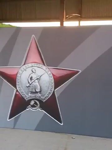 The video shows a close-up view of a wall mural featuring Soviet-era military insignia. On the left, a large red star with a silver outline is visible. To its right, a medal for the Defense of Leningrad hangs from a green and white ribbon. The medal is circular and displays figures of soldiers and a lighthouse. Further to the right, a circular emblem with the hammer and sickle and the text "PROLETARIANS OF ALL COUNTRIES, UNITE!" is prominently featured. The background of the mural is a gradient of gray with diagonal stripes. The mural appears to be indoors, possibly in a hangar or exhibition space, with a wooden ceiling structure visible at the top of the frame. The lighting is bright, suggesting daytime or artificial illumination. The camera pans slowly across the mural, providing a detailed view of each element.