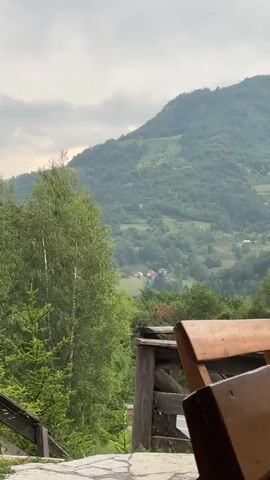 This media file shows a scenic view of a mountainous landscape, likely Mekića Kula in Montenegro. The foreground features a wooden bench and railing, suggesting a viewpoint or seating area. Behind this, lush green trees, including a prominent birch tree, dominate the left side of the frame.

The midground reveals a valley dotted with several houses with red roofs, nestled amongst rolling hills covered in dense green foliage. The background is dominated by a large, verdant mountain that slopes upwards, disappearing into a cloudy sky. The time of day appears to be overcast, possibly late afternoon or early morning, given the diffused light. The weather seems to be cloudy or possibly humid, contributing to the soft, hazy appearance of the distant mountains.

No people are visible in the media. The overall mood is tranquil and serene, highlighting the natural beauty of the mountainous region. The presence of the bench implies a moment of rest or observation, inviting the viewer to appreciate the expansive vista. No text is visible.