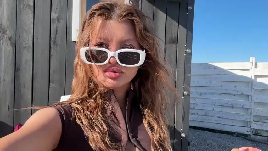 A woman with long blonde hair and large white sunglasses is taking a selfie outside on a sunny day. She's wearing a brown sleeveless top and pouting her lips at the camera.  Behind her is a gray wooden wall and a white picket fence. She puts her hand up to adjust her hair.