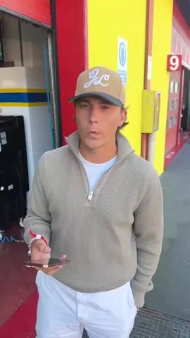 The video opens on a man wearing a tan baseball cap and a neutral-colored quarter-zip sweater over a white t-shirt. He has white pants on and is holding a cell phone in his left hand, along with what appears to be a tool. He is standing in what looks like a garage or workshop area. The background is a busy scene with several people working on bright green race cars. The cars have yellow and blue racing stripes and appear to be Lamborghinis. Some car hoods are open, and mechanics are seen attending to the engines. One car has the number 19 on it. The garage has red and yellow walls and a large open garage door in the background. In the foreground, a green car part, possibly a spoiler or fender, with blue graphics is lying on the ground. The video then cuts to different angles of the garage and the race cars, with the man in the foreground occasionally visible in some shots.