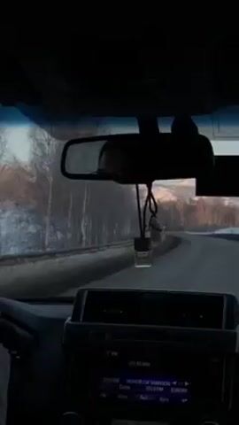 A car is driving on a road next to a snow-covered forest. The road appears to be in good condition, and the sky is lightly overcast. In the distance, snow-covered mountains can be seen. The car's dashboard is visible, with a radio display showing various stations. An air freshener hangs from the rearview mirror. The overall scene suggests a winter road trip through a mountainous region.