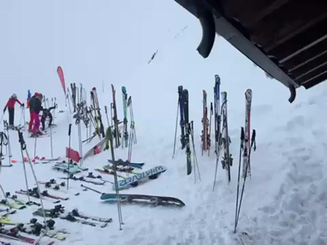 The video shows a snowy mountain scene, likely at a ski resort. Numerous skis and ski poles are stuck in the snow near what appears to be a building with a covered area. In the background, a group of people are skiing or snowboarding on a slope. The weather is overcast, with limited visibility. A young man is briefly seen on a deck looking out at the scene, and there are several flags visible behind him.  The flags appear to be French and Swiss. Snow is actively falling.