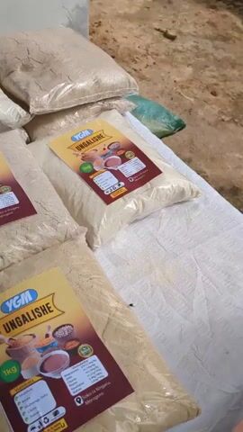 Here is a concise description of the scene:

The video shows an outdoor setting with a variety of bagged food products arranged on a table covered with a white cloth. The products appear to be different types of flour and beans. Several bags are labeled "YGM UNGALISHE 1kg" along with nutritional information. Other bags contain ingredients like "Nafaka." A white plastic chair is positioned near the table, holding a denim jacket and a black handbag. Two black notebooks lie on the table alongside the food items. The camera pans across the arrangement, providing a view of the various products and the surrounding environment. The scene suggests a possible market display or storage area for food items.