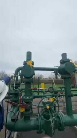 A person in a hard hat and blue jacket stands near a series of green pipes and valves, labeled with yellow tags. The person is operating a valve, releasing a hissing gas or vapor into the air. The sky is overcast. In the background is a green field and some trees.