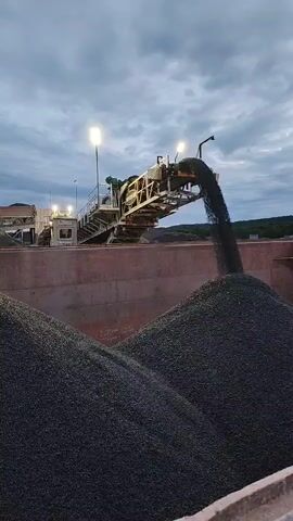 A conveyor belt system is actively dumping a dark, granular material, likely coal or aggregate, into a large barge or industrial bin. The material cascades from the end of the conveyor, forming a substantial pile. The scene takes place outdoors under a cloudy sky with artificial lights illuminating the conveyor. The background suggests an industrial or port environment with some trees and structures visible in the distance. The conveyor system is elevated and appears to be part of a larger operation.
