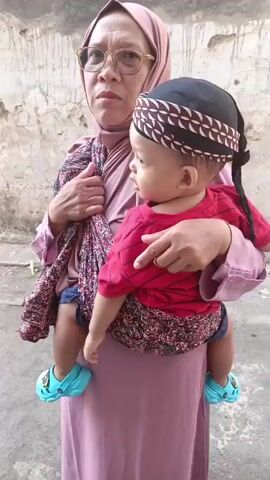 A woman wearing a pink hijab and glasses is holding a baby wrapped in a patterned cloth sling. The baby is wearing a red shirt and a black head covering with a white and brown pattern. The baby is facing to the right, looking away from the camera. The woman is looking at the camera. The background appears to be an outdoor setting with a textured wall. The baby is wearing blue clogs.