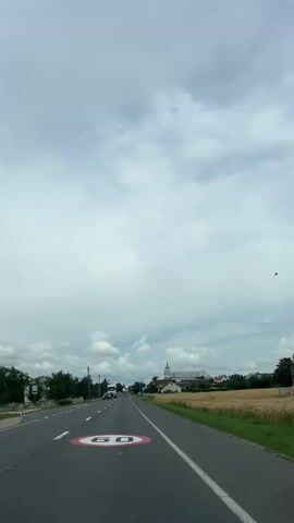 The video shows a view from a car driving down a road. A speed limit sign of 60 is visible on the road in the foreground. On the left side of the road, there are trees and some buildings. On the right side of the road, there is a field of grain. In the distance, there is a town with a church. Several cars and a truck are also visible on the road. The sky is cloudy.