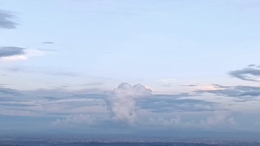 The video shows a wide shot of the sky and a distant landscape. The sky is predominantly light blue with scattered white clouds of varying shapes and sizes. A distinctive, tall, puffy cloud formation dominates the center of the view. The landscape below is faintly visible, with an indistinct urban area appearing in the distance, suggesting an elevated viewpoint.  The overall tone is serene and atmospheric.