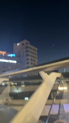 The video pans across a building at night in Nicosia, Cyprus. The building features a prominent sign that reads "AMERICAN COLLEGE" in illuminated white letters. Above this, on the rooftop, are decorative structures resembling large umbrellas or canopies, illuminated with warm yellow and orange lights. To the left of these structures, a section of the rooftop is bathed in a vibrant, glowing blue light, possibly indicating a pool or lounge area. The sky is a deep, dark blue, suggesting nighttime. A few lights are visible in the windows of the building, and some air conditioning units are mounted below the windows. In the distance, a faint, small light can be seen in the sky, which might be a star or a distant light source. The foreground shows part of a modern structure with a white railing and glass, suggesting the viewpoint is from a balcony, walkway, or a vehicle with large windows. The overall scene conveys an urban nighttime ambiance, with the illuminated building and rooftop features standing out against the dark sky.