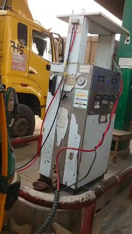 The video shows a man standing at a gas station next to a filling station. He's wearing a black shirt and gray pants and has money in his hand. The gas station is a large, somewhat dilapidated-looking piece of equipment with a variety of gauges and connections. It is white with a stainless steel front. There are various vehicles in the background, including a yellow truck and a trailer. The station is beneath a corrugated steel roof.