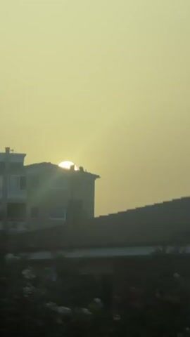 The video displays a hazy yellow sky with the sun visible behind a building. The camera pans across rooftops with chimneys and then focuses on a body of water that reflects the hazy sky and the surrounding trees.