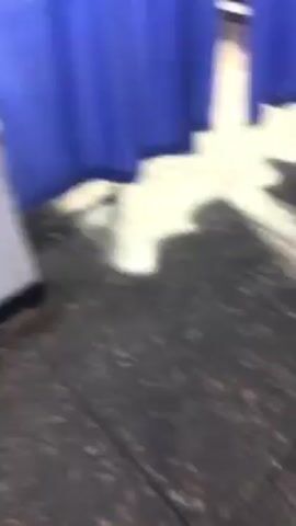The video shows a sparsely lit corridor. On one side are blue curtains hanging from white supports. The floor is speckled. There are doors and a picture on the wall further down the corridor. Towards the end of the video, the camera focuses on a gray wall.