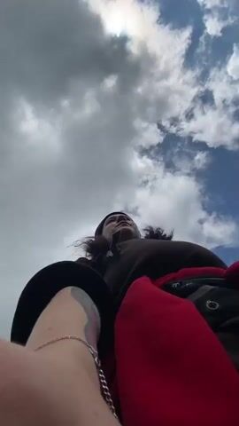 A low-angle, first-person perspective video shows a person with dark, wavy hair, wearing a black beanie, a dark brown long-sleeved shirt, red pants, and a silver bracelet. They are holding a phone or camera in their left hand, taking a selfie. The person is outdoors, in a grassy, hilly landscape under a cloudy sky. In the background, other people can be seen walking along a path in a field. The video shows the person turning their head from side to side and looking towards the camera, smiling. The environment is natural and rural, with rolling hills and green grass.