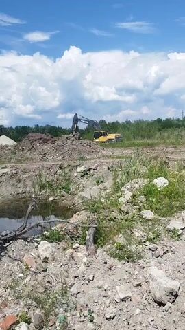 This media file captures a view of a construction site under a bright blue sky with fluffy white clouds. In the background, a yellow Volvo excavator is actively working on a large pile of earth and debris. The excavator's arm is raised, suggesting it is in the process of digging or moving material. The foreground is a rough, uneven terrain covered in rocks, dirt, and some sparse green vegetation. A small pool of water with a fallen tree branch lies in the mid-ground. The overall scene depicts ongoing construction or excavation work in a rural or semi-rural outdoor setting. The lighting indicates it is daytime, likely midday, given the bright sunlight. The weather appears clear and pleasant. While no people are visible, the presence of heavy machinery clearly indicates human activity. The location context provided is Rozdil, Ukraine.