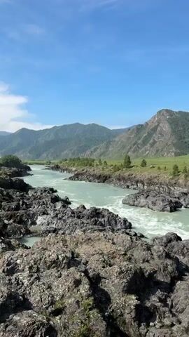 The video begins with a view of a wide, turbulent turquoise river flowing through a rocky gorge. Jagged dark rocks line the riverbanks, with lush green hills and mountains in the background under a bright blue sky with scattered clouds. The camera then pans to the right, revealing a suspension bridge. The bridge has a red and white railing and appears to be made of metal. Thick steel cables are visible, with visible rust and wear on the hardware. As the camera moves across the bridge, more of the mountainous landscape and the river below can be seen. The bridge deck is rough and appears to be made of asphalt or packed dirt. There are signs on the bridge, including one with the number 3.5, possibly indicating a weight limit. Graffiti is visible on a concrete structure at the edge of the bridge. The camera continues to pan, showing more of the detailed structure of the bridge and the natural scenery.