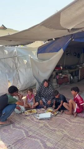 The video shows five people sitting on a woven mat outdoors under a makeshift shelter made of tarps. In the center, an older woman wearing a headscarf and patterned dress sits cross-legged. To her left, a young girl in a pink tank top sits similarly. To her right, two boys are seated, one on each side. The boy closer to the woman is wearing a dark t-shirt and is actively eating from a plate with a utensil. The boy further to the right wears a maroon shirt with white accents and is also seated cross-legged. On the far left, a young man in a dark t-shirt and blue jeans is crouched down, appearing to serve food or interacting with one of the children. In front of the group, several plates of food and a white foil-wrapped container are arranged on the mat. Behind the seated individuals, the shelter's interior is visible, showing kitchen items and storage containers. The overall environment appears to be a simple, possibly temporary dwelling in a sunny, outdoor setting.