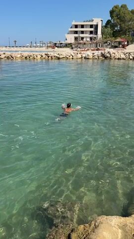 A person is swimming in clear, shallow turquoise water. The person is wearing dark shorts and a dark swim cap. The person's head is periodically submerged in the water as they swim. In the background, there is a rocky shoreline with a modern, multi-story building situated on the land behind it. There are also some trees and beach chairs visible. The sky is clear and blue, indicating a sunny day. The water is calm with small ripples.