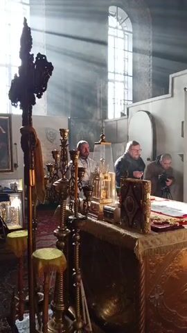 The video shows an indoor scene inside a church, with sunlight streaming in through a large arched window. Three men are standing around a table covered in a golden cloth. There's a priest in white robes standing closest to the camera and blessing the table, a man in a black jacket holding a chain with small crosses, and another man setting up a camera on a tripod. The table is adorned with religious artifacts including candlesticks, a decorative book, a reliquary in a glass case, and a cloth with crosses laid out on it. To the left of the table, there's a large ornate cross and a painting visible in the background. The atmosphere seems reverent.