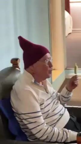 An elderly man sits in a leather armchair, wearing a purple pointed hat, glasses, and a white-and-blue striped shirt. He's holding a small green object that resembles a popsicle or a small candle. The man is looking towards the right, and appears to be speaking.  Behind him is a pale blue wall, and an open doorway leading into another room. A bed can be seen in the background.