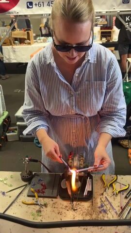 A woman in a blue and white striped shirt and sunglasses is using a torch to melt and shape glass rods on a table. The torch is mounted on a small apparatus with red and yellow fittings. Various tools are scattered around the workspace, including pliers and other small instruments. There are also finished glass pieces, some shaped like flowers, on a black surface. The woman is focused on her work, carefully manipulating the glass in the flame. The background shows a busy indoor market scene with other stalls and people walking around. The environment appears to be a crafts fair or similar event.