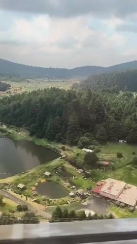The video shows an aerial view of a landscape featuring lakes, forests, and buildings, captured from what appears to be a moving train or vehicle. The sky is overcast. The landscape consists of rolling hills covered in dense green forests with a valley visible in the distance. In the foreground, there are multiple lakes or ponds, some of which are surrounded by small buildings and structures, possibly cabins or recreational facilities. The presence of a metal rail or barrier along the bottom of the frame confirms that the video is taken from inside a moving object.